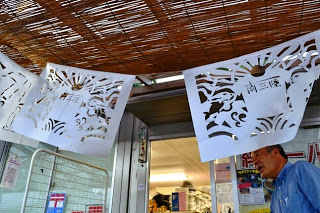 男性が立っている商店街の入り口の上に、南三陸の文字とともに魚が描かれた切り絵が飾られた写真