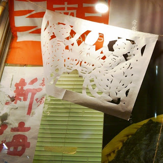 窓ガラスの前に子供と竜のような動物がデザインされた切り絵が吊られている夜の店舗の写真