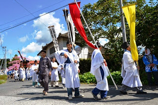 青空の下、白装束や和装の参加者たちが赤黄の旗を掲げながら道路を練り歩く行列の写真
