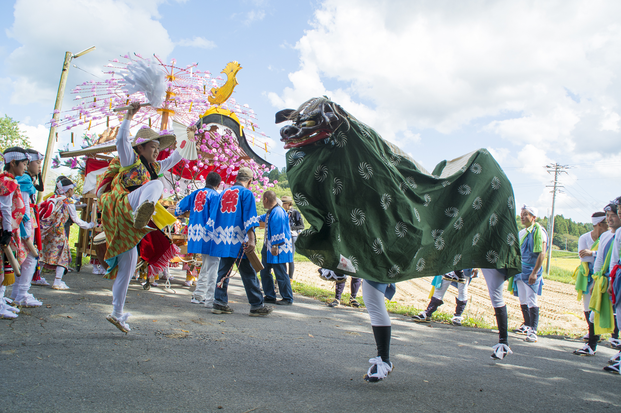 青空の下で獅子舞と扇を持つ踊り手が向かい合い、背後に華やかな山車と法被姿の人々が並んでいる写真
