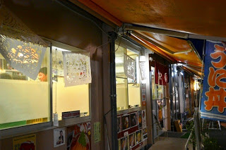 紙を切り抜いたきりこの飾りが商店の窓に貼られており、店内では人が調理をしている様子が見える夜の商店街の写真