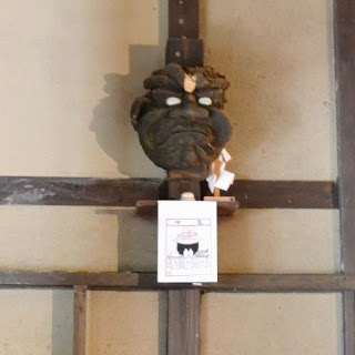 木の壁に取り付けられた黒い鬼のような面の下に、釜のイラストが描かれた紙が掲げられているカマガミサマの写真