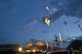 屋台や人々が集まる夏祭り会場の薄暗くなった夕暮れの空に、カラフルな花火が打ち上がっている様子の写真