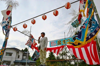 提灯や旗でにぎやかに飾られた櫓の上で、男性がマイクを持って開会の挨拶をしている盆まつりの写真
