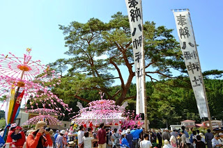 大きな一本松の下、巨大なピンクの花傘を囲むようにして多くの人々が集まり賑わっている写真