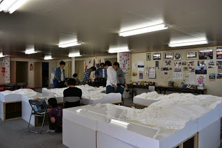 壁に沢山の写真が飾られた室内に白い立体地形模型が複数並び、複数の人たちがその模型を囲んで話し合っている写真