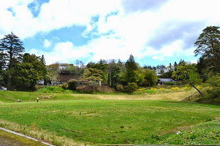 広々とした草原の奥に木々に囲まれた茅葺き屋根の古民家や建物が建ち、のどかな山里の風景が広がっている、ひころの里の全景写真