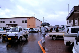 雨が降る中、駐車場や建物の前に沢山の車が停まっている間を、傘をさして建物へ向かって歩いている男性の後ろ姿の写真