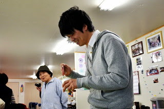 背景に年配の女性が笑顔で立っている明るい室内で、若い男性が笑顔で小さな透明の旗を手にしている写真