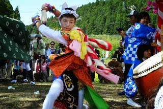 周囲の人々が見守る中、太鼓の音色に合わせて鮮やかな衣装を身にまとった少年が腕を大きく振って舞を踊っている写真