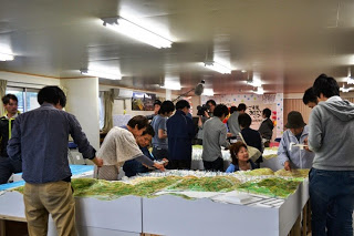 多くの人が立体模型の周囲に集まり、奥に設置された模型の傍で、ガンマイクやENGカメラでテレビ局が撮影を行っている様子の写真