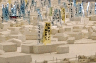 白い地形模型の中に家や建物が配置され、青や黄色の短冊に地名や人名が書かれて多数立っている様子をアップで撮影した写真