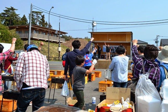 屋外ステージのような場所に立つ人々が拳を掲げている広場で、間隔をあけて設置されたバーベキューコンロの周りにいる人々も一緒に拳を掲げている祭りの様子の写真