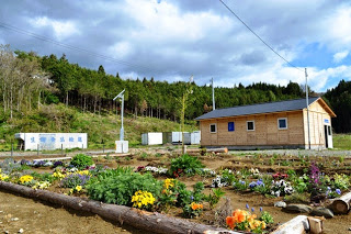 花壇に色とりどりの花が咲き、その奥に木造の小さな建物と森が広がっている写真