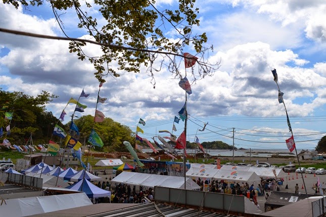 雲が浮かぶ青空の下、こいのぼりや大漁旗が空にたなびき、沢山の屋台と人でにぎわう港近くの祭り会場全景の写真