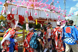 子どもたちが華やかな衣装を着て、紅白幕とピンクの花飾りで装飾された山車を運んでいる様子の写真
