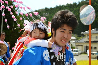 笑顔の青い法被を着た男性が、嬉しそうに笑っている小さな子をおんぶしている祭りの一コマの写真