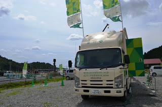 仙台銀行のロゴが入った移動型店舗車に緑と白の旗が上がっており、正面を向いて停車している写真