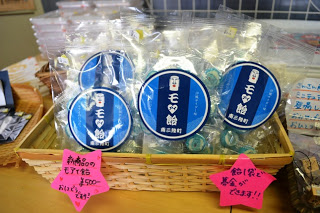 「モ飴」と書かれた青い丸いラベルが付いた飴の袋がかごに並べられ、価格表示の手書きポップも見える写真