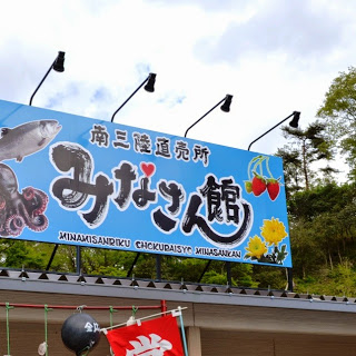 魚やタコ、サクランボ、花などのイラストが描かれている、「南三陸直売所 みなさん館」と書かれた青い看板の写真