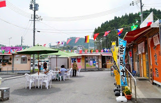 色とりどりの国旗が張られた屋外スペースに、プラスチックのテーブルと椅子が置かれ、数人が座っている商店街の広場の写真