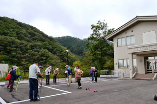 背景に緑の山々を望む中、白い建物の前の広い駐車場で、トレッキングポールを持った十数人の方々が準備運動をしている様子の写真