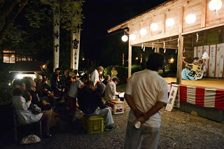 夜の神社境内で、提灯に照らされた舞台を前に椅子に座って神楽を鑑賞する人々の写真