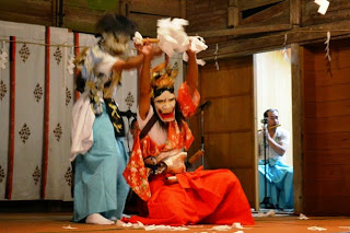 能舞台の上で、鬼のような面をかぶった水色と赤色の衣装を着た二人の人物が紙を舞わせながら演舞している写真