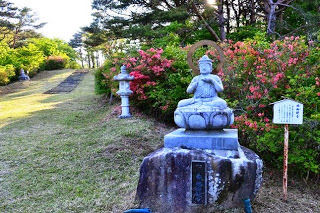 つつじの咲く小道の脇に石仏や石灯籠が並び、整備された芝生の斜面が奥へ続いている写真