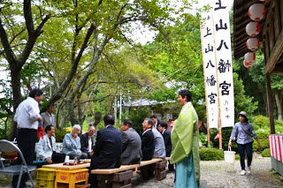 神社の境内に集まった男性たちが簡易ベンチに座って会話をし、傍で黄緑色の着物を着た神主さんが立って見ている写真
