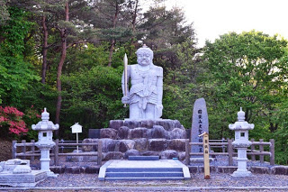 周囲を木々や草木が囲む屋外に、柵の外にある左側の台の上に倒壊した両足が置かれ、両脇には石灯籠があり、柵の中の右側には背の高い石碑が立ち、石の台座の上に、剣を持った足の無い大聖不動明王が立っている写真