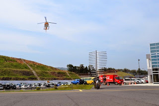 病院の前の駐車場上空でオレンジ色のヘリコプターがゆっくりと着陸体勢で降下している写真