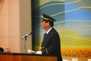 制服と制帽を着用した佐藤町長がマイクの前でスピーチしている出初め式の式典の様子の写真