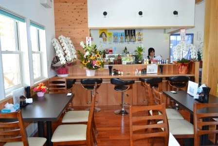 花が飾られたカウンターの前に椅子が並び、その周りにテーブル席が配置され、奥にはスタッフが見えるカフェ店内の写真