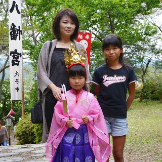 石階段を上がった神社ののぼり旗の前で、ピンクと紫の衣装を着た女の子と母親、姉らしき人物が並んで写っている写真