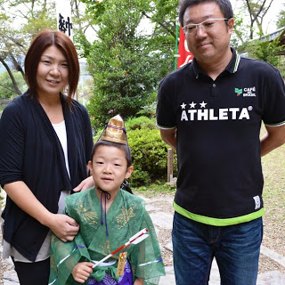 緑色の装束を着た男の子が破魔矢を手に持ち、両脇に立つ両親とともに笑顔で写っている写真