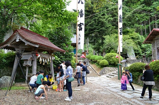 神社の境内で手水舎の水を使って子どもたちが手を清め、大人たちが見守る中、石畳の参道をピンクの衣装を着た女の子が母親と一緒に撮影している写真