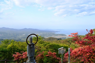 赤く色づいた木々の傍に、遠くの町と海が広がる風景を見下ろすように立っている観音菩薩の石像の後ろ姿の写真