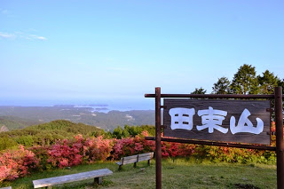 ピンク色のつつじが咲いている山頂に「田束山」と大きく書かれた木の看板が立ち、眼下に緑の山々と森、海が広がっている風景の写真