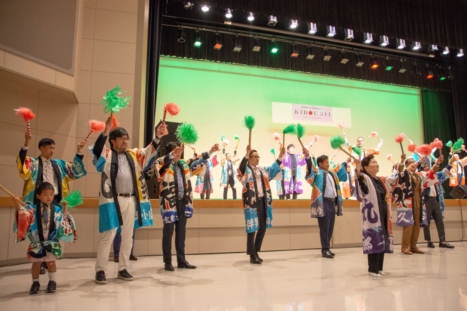 背景は緑色の照明で照らされ、後方の壁には横断幕が掲げられているステージ上とその前で、大漁旗柄の法被を着た人々が並び、カラフルなポンポンを使ってパフォーマンスをしている写真