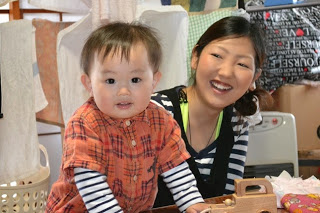 赤いチェック柄の服を着た小さな子どもがテーブルの上に手を置いて立ち、後ろでボーダー柄のシャツの女性が笑顔で見守っている写真