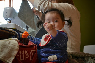 ミシンで布を縫っている女性の傍で、小さな女の子がカメラに向かって手を伸ばしている写真