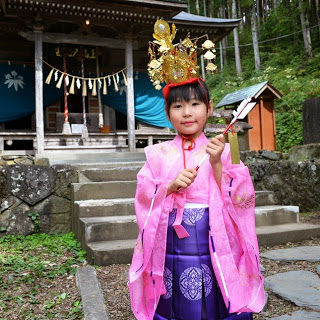 拝殿の前に立ち、金の冠とピンクの衣装を身につけた女の子が両手で破魔矢を持ち笑顔でポーズをとっている写真。