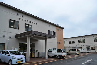 白とベージュの二階建ての建物が並んでいて、前には軽自動車が数台駐車しており、正面入口には屋根がついていて、建物の壁には「公立南三陸診療所」と書かれている診療所の写真