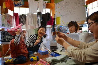 室内に洗濯物が干され、ミシンが置かれた部屋で、数人の大人が手芸を楽しんでいて、中央の女性がピンク色の布を被った小さな子どもに向かって人差し指を上げたポーズをしている写真