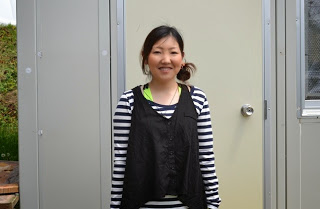 明るい外で、白と黒のボーダー柄のシャツを着た女性がプレハブ建物のドアの前で笑顔で立っている写真