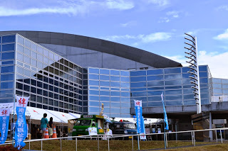 近代的なガラス張りの建物の前に屋台やのぼり旗が立ち、来場者が中に入っていく様子が見える写真