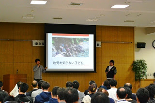 「地元を知らない子ども」と書かれたスライドを背景に、二人の発表者が前に立って話をしており、聴衆が真剣に聞いている写真