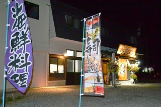 夜の店先に「海鮮料理」や「寿司」と書かれたのぼり旗が立ち、入り口上の看板や建物に明かりが灯っている開店中の寿司店の外観の写真