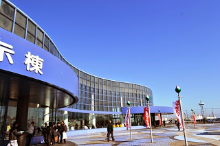 大きくカーブしたガラス張りの近代的な夢メッセみやぎの建物の入口前で、複数の来場者が青空の下を歩いており、赤いのぼり旗が並んで風に揺れている様子を写した展示会場入口の写真